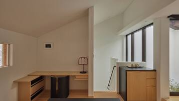 A modern, minimalist room with a wooden desk, a black chair, a small lamp, and a view through large windows.