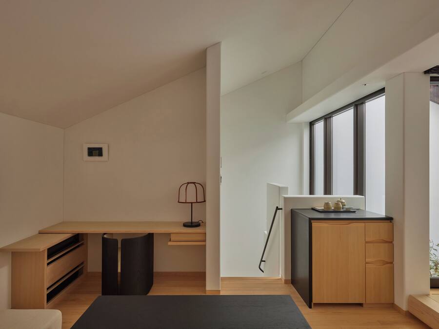 A modern, minimalist room with a wooden desk, a black chair, a small lamp, and a view through large windows.