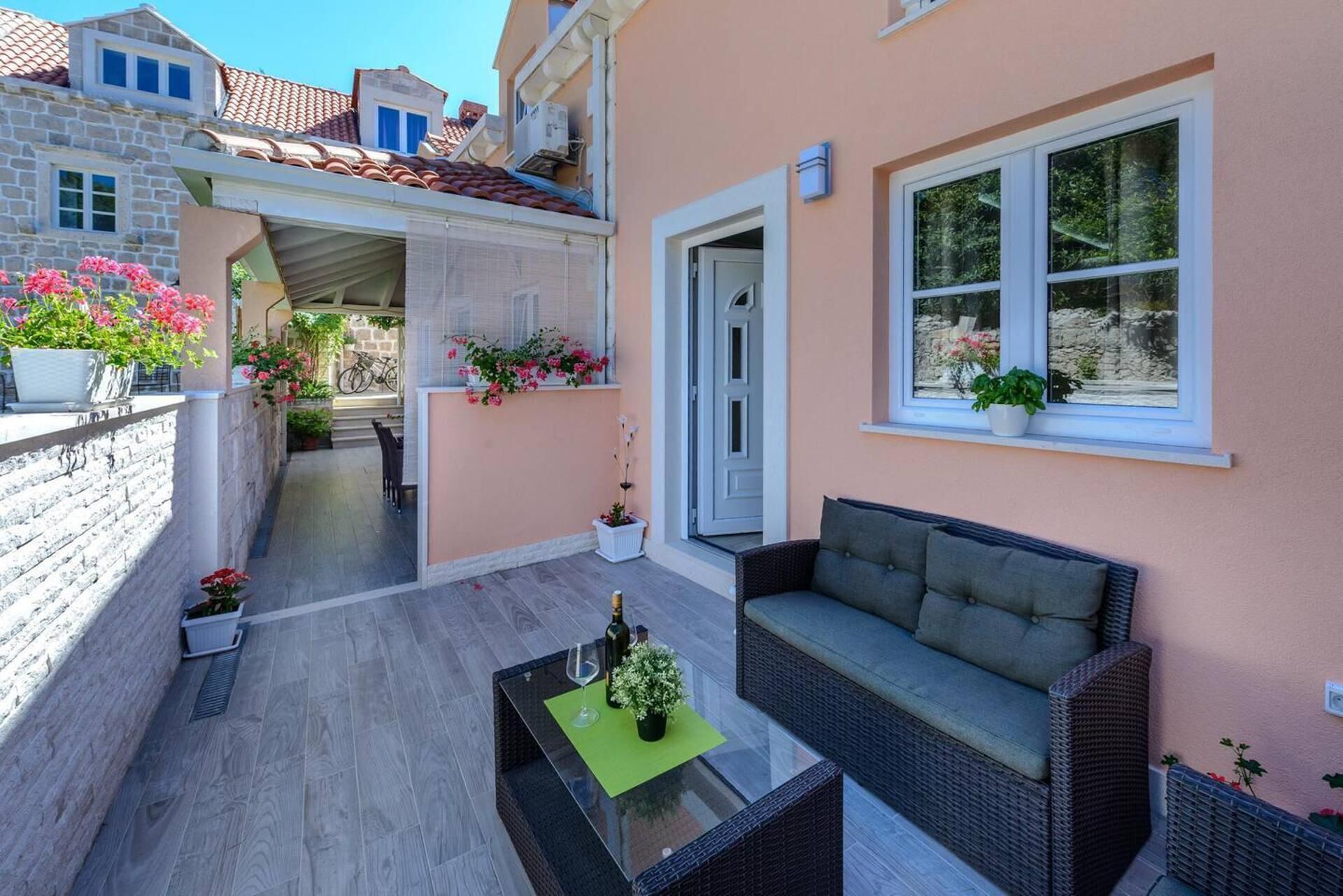 A balcony with a wicker sofa, a glass table with a wine bottle, and potted plants.