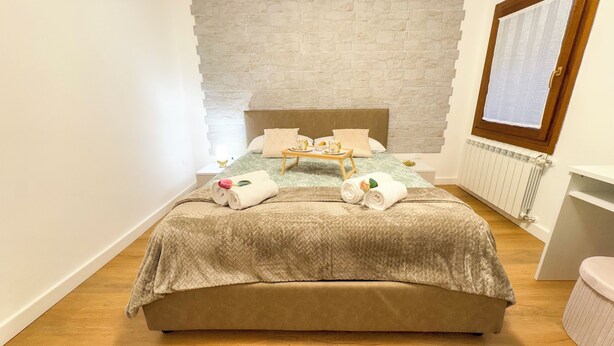 A neatly made bed with a herringbone-patterned bedspread, a wooden bedside table with a tray of breakfast items, and a stone-textured wall behind the bed. - C A L I by Ca-sa Check (Chioggia)