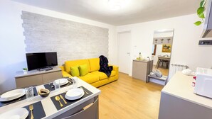 A modern living room with a yellow sofa, a flat-screen TV, and a dining area with a table set for two. - C A L I by Ca-sa Check (Chioggia)