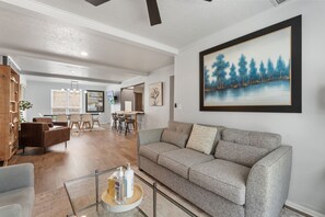 A spacious living area with a grey sofa, wooden furniture, and a large painting of a forest scene. - Spacious Retreat 8 Bedroom River (Bethany)