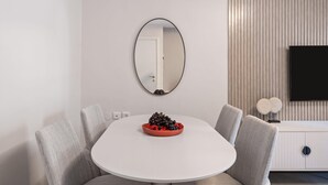 A modern dining area with a white table, grey upholstered chairs, a bowl of fruit, a round mirror, and a flat-screen TV on a white cabinet. - HaHavatselet 19 - Isrentals - Superior 5 (Jerusalem)