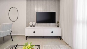 A modern living room with a flat-screen TV mounted on a white cabinet, a grey armchair, and a glass coffee table with decorative items. - HaHavatselet 19 - Isrentals - Superior 5 (Jerusalem)