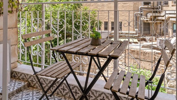 A balcony with a wooden table and chairs, a patterned floor, and a view of a building with multiple windows. - HaHavatselet 19 - Isrentals - Superior 5 (Jerusalem)