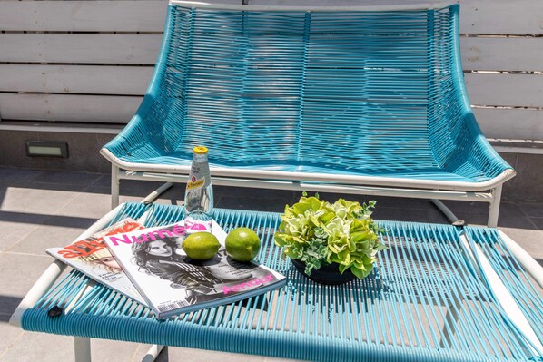 Una silla de mimbre turquesa con una revista, dos limones y una pequeña planta en maceta sobre una mesa de metal.