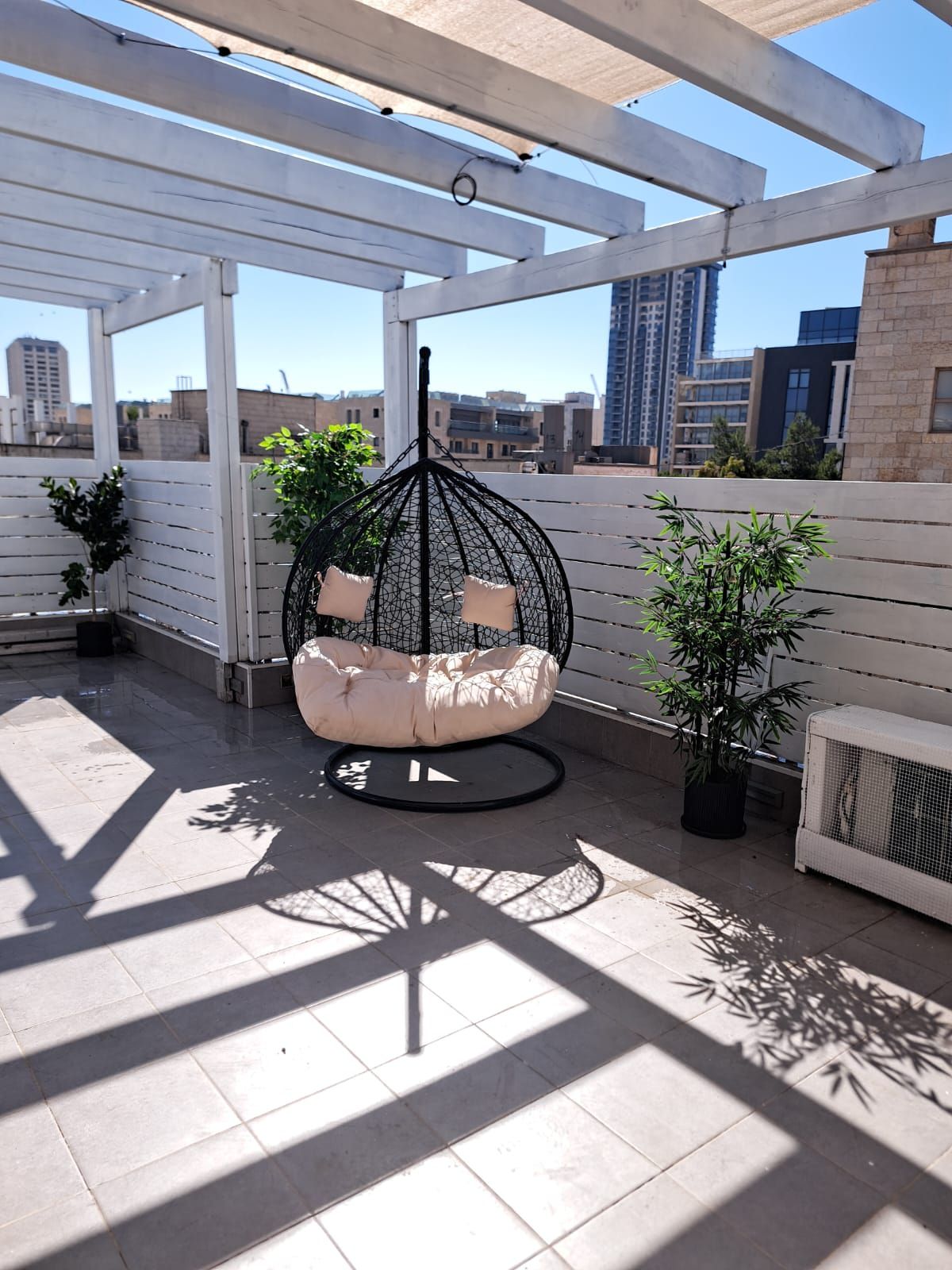 A rooftop patio with a hanging chair, potted plants, and a view of the cityscape.