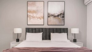 A neatly made bed with a grey headboard, two bedside tables with lamps, and two abstract paintings on the wall.