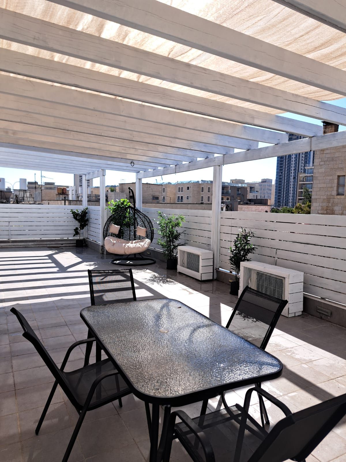 An outdoor patio with a table and chairs, a hanging chair, potted plants, and air conditioning units.