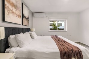 A modern bedroom with a large bed, two wall-mounted air conditioners, and a window with a view of buildings and trees.