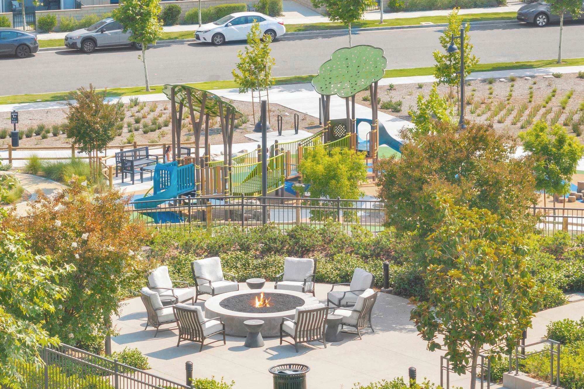 Un parque infantil con toboganes y estructuras para escalar, rodeado de árboles y una zona con hogar para fogata y asientos.