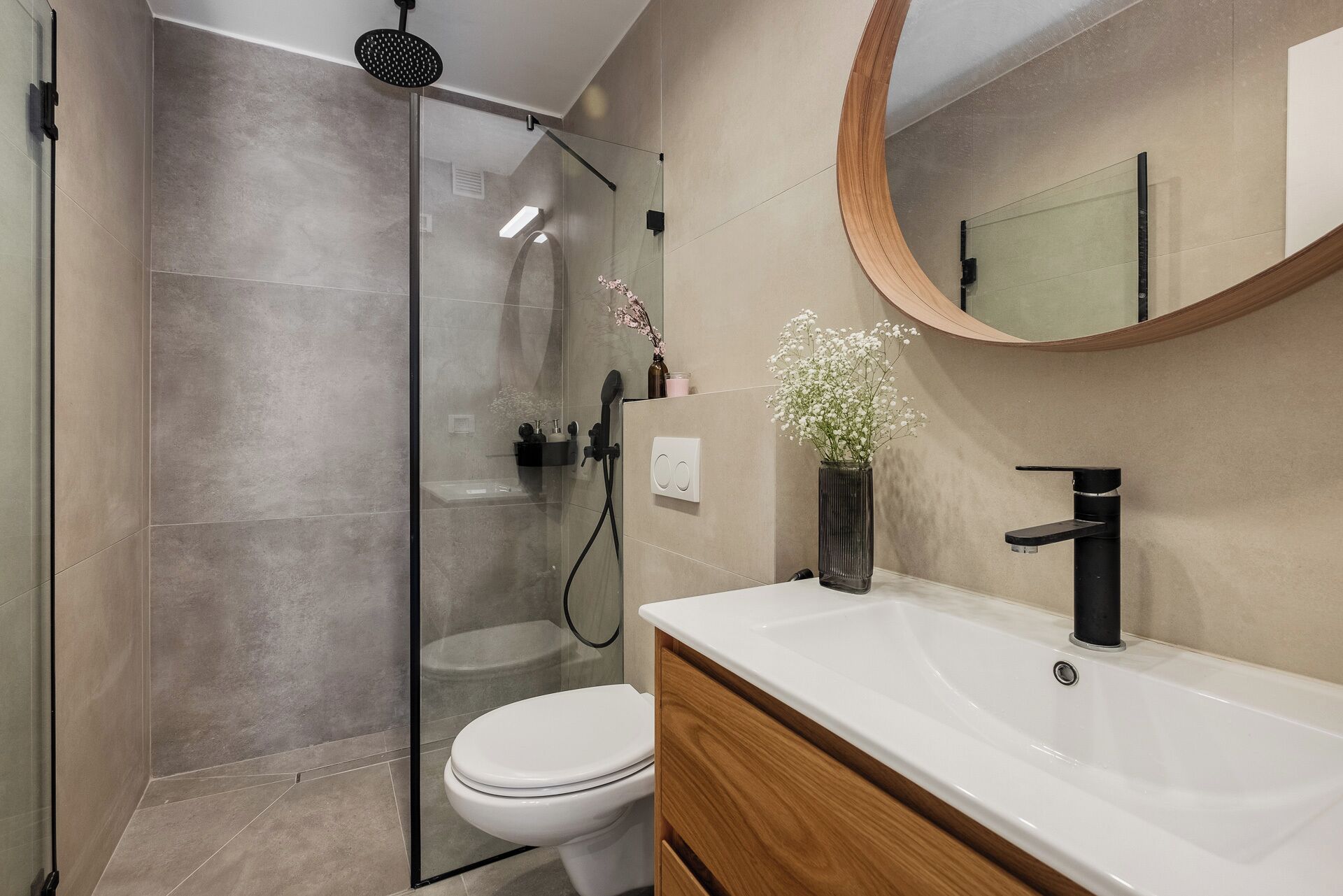 A modern bathroom with a glass shower enclosure, a white toilet, a wooden vanity, and a round mirror.