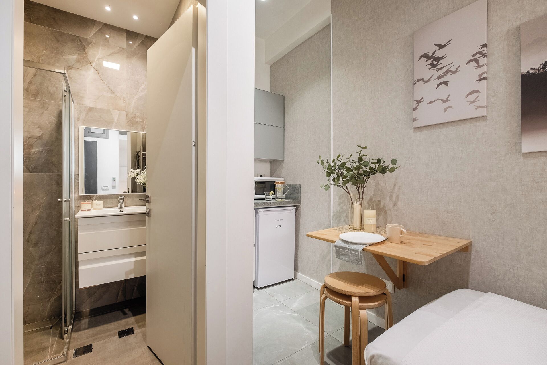 A modern bathroom with a glass shower enclosure, a vanity with a mirror, and a small seating area with a table and a stool.
