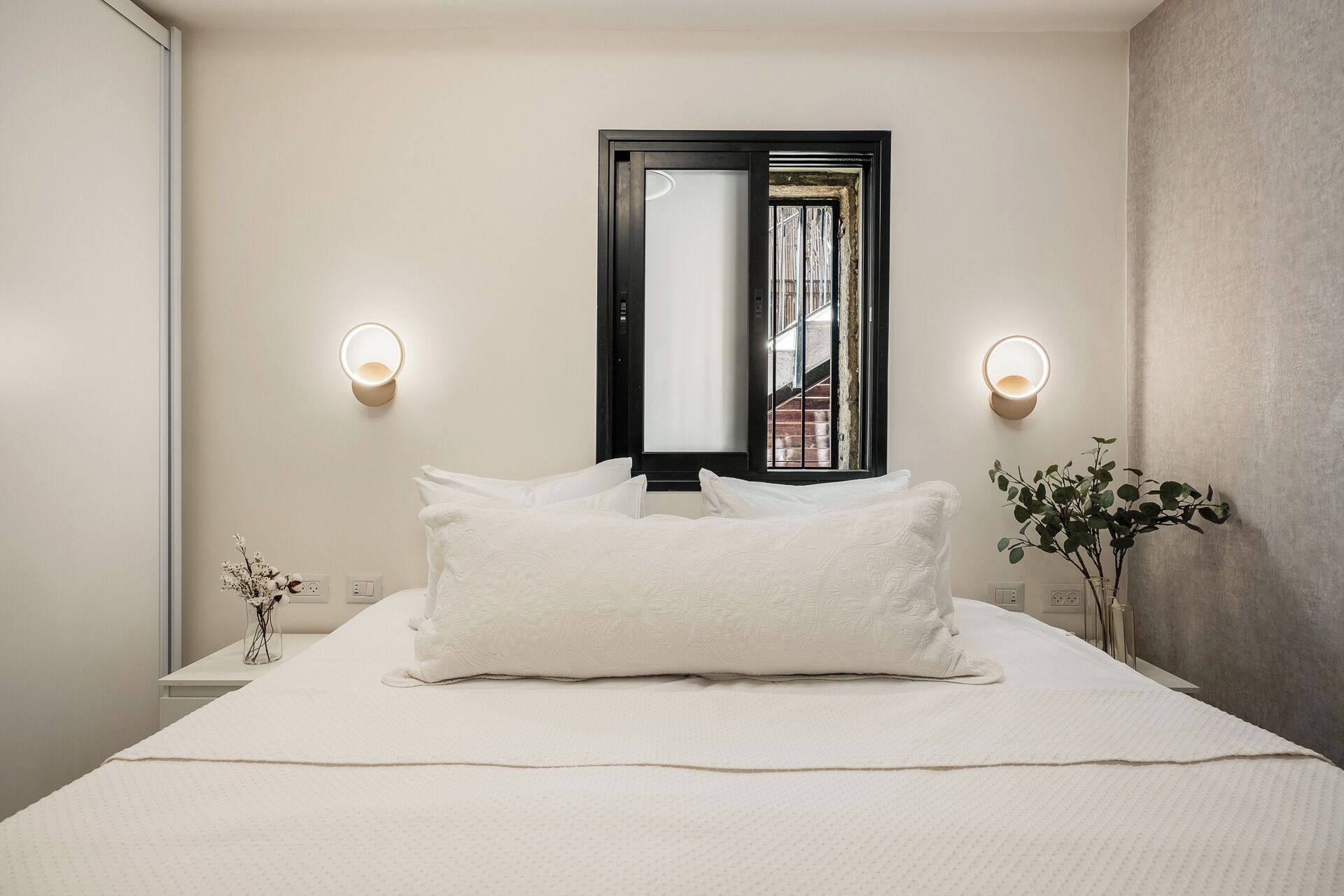 A neatly made bed with white linens, two bedside tables with lamps, a mirror above the bed, and a potted plant on the right side.