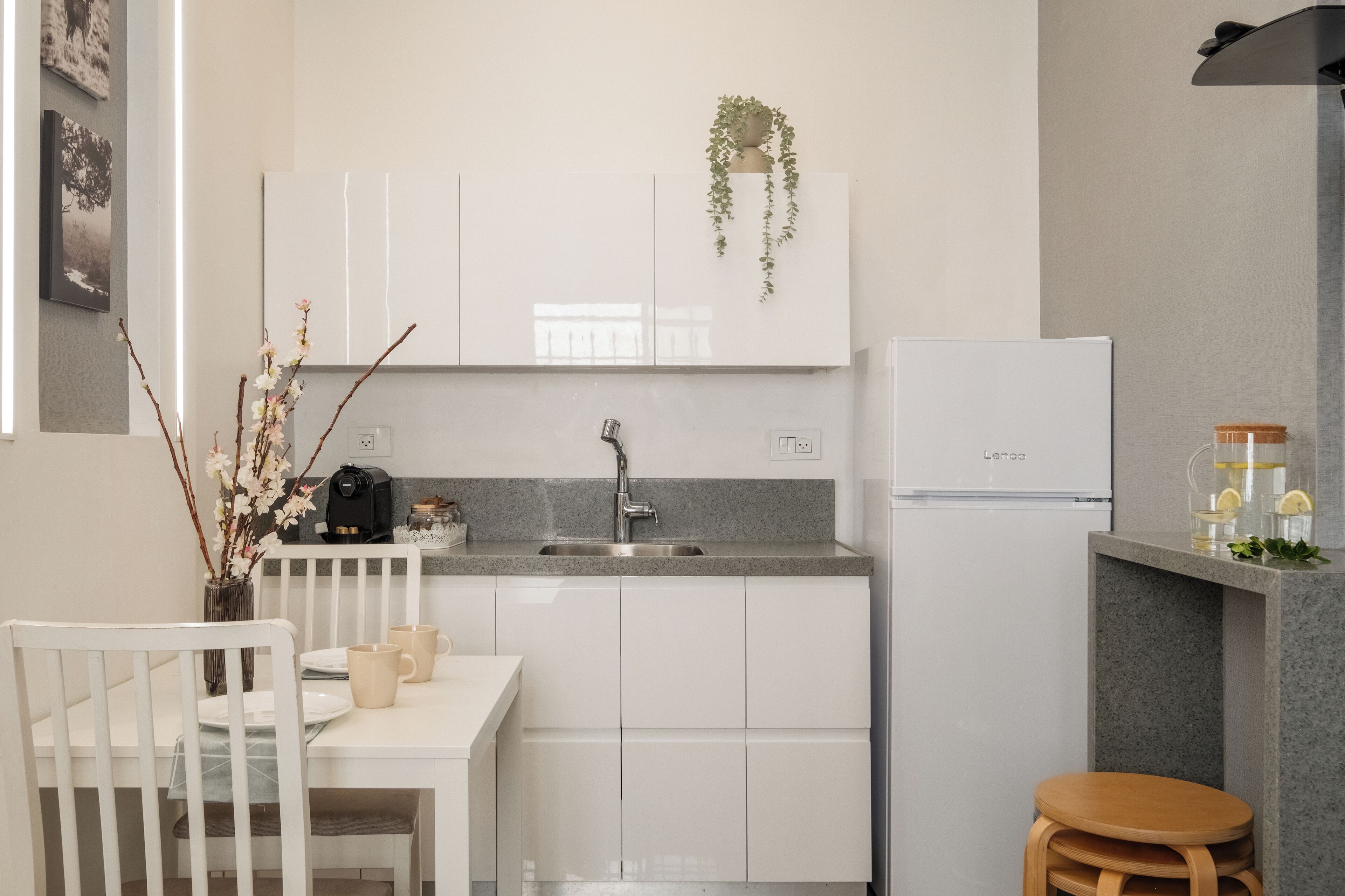 Une cuisine moderne avec des meubles blancs, un plan de travail en granit, un évier et un réfrigérateur. On y trouve une petite table à manger avec des chaises et un vase décoré de branches.