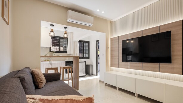 A modern living room with a large flat-screen TV mounted on the wall, a dark sofa, and a kitchen area visible through an open door. - baraca boutique (Jerusalem)