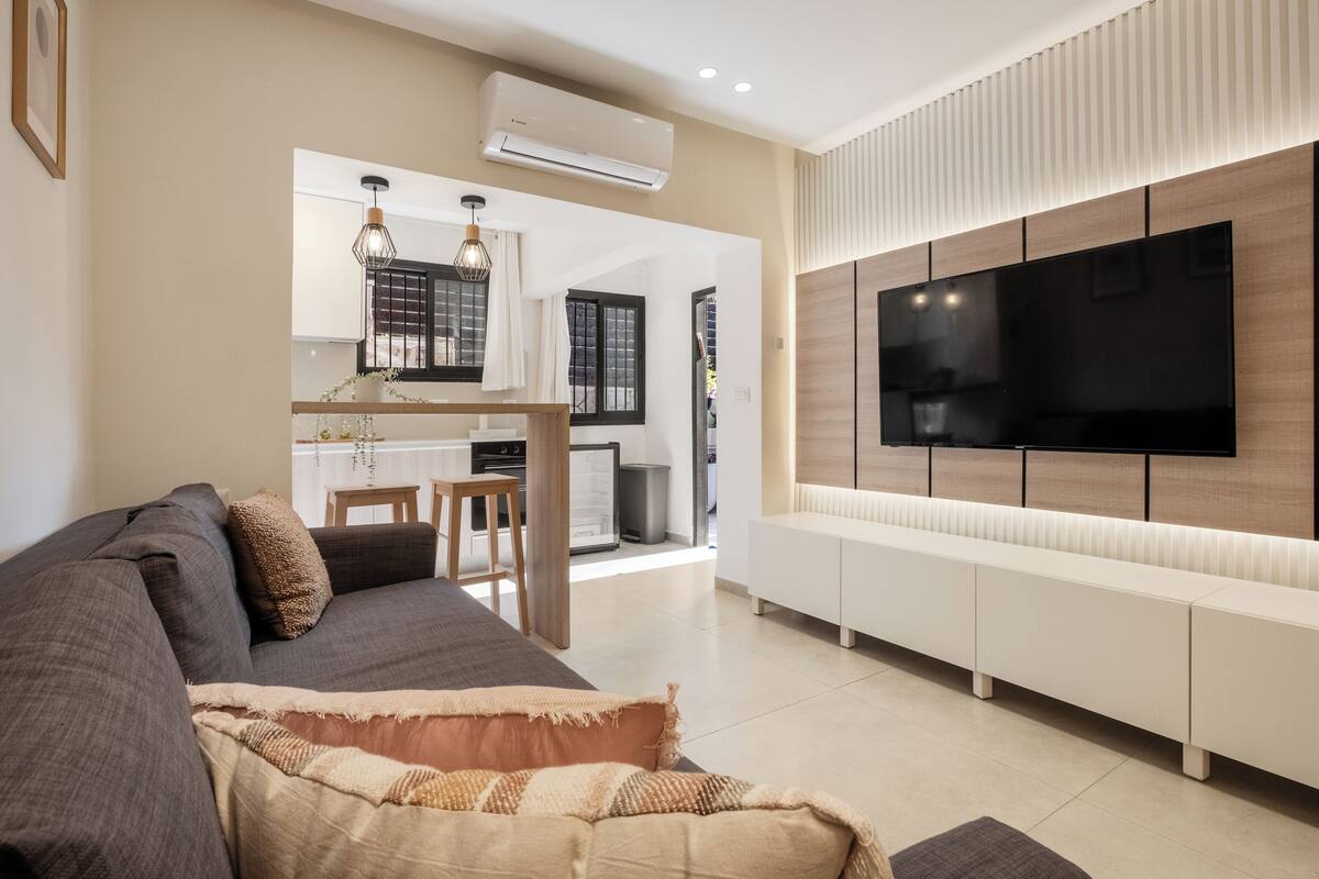 Uma sala de estar moderna com uma grande TV de tela plana na parede, um sofá escuro e uma cozinha visível através de uma porta aberta.