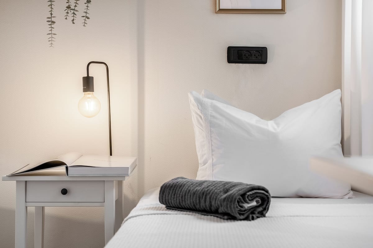 Cama bem arrumada com travesseiro branco e manta cinza enrolada, mesa de cabeceira com um livro aberto e luminária fixa na parede.
