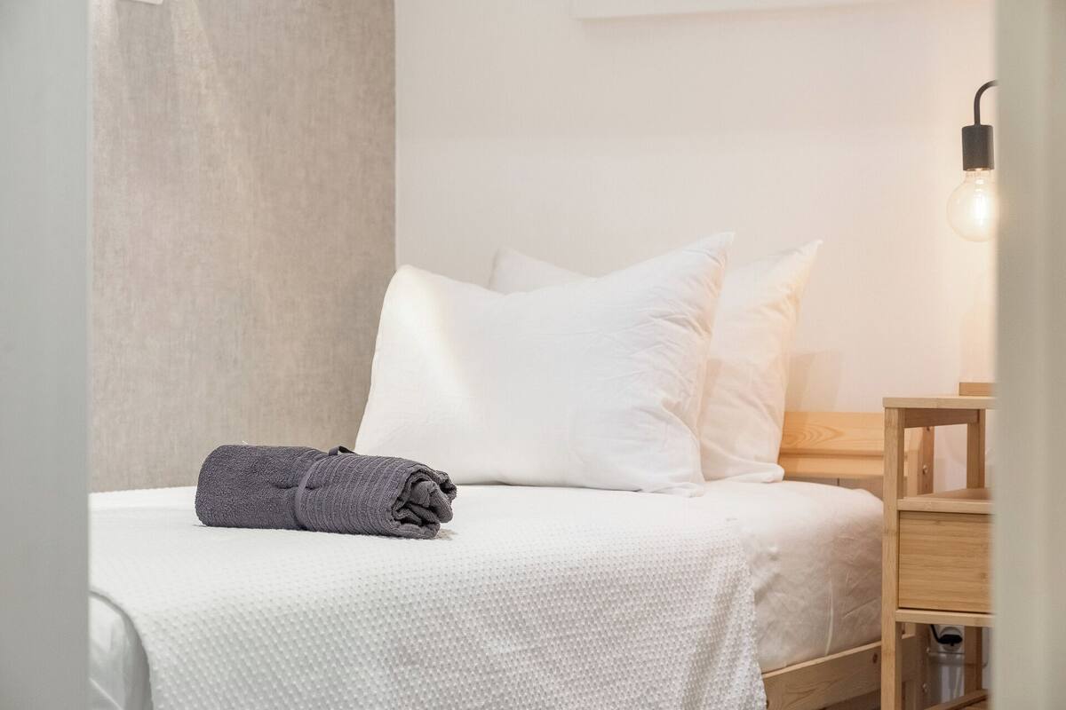 Cama bem arrumada com lençóis brancos e uma toalha cinza dobrada, mesa de cabeceira de madeira com uma luminária e um móvel com gavetas de madeira.