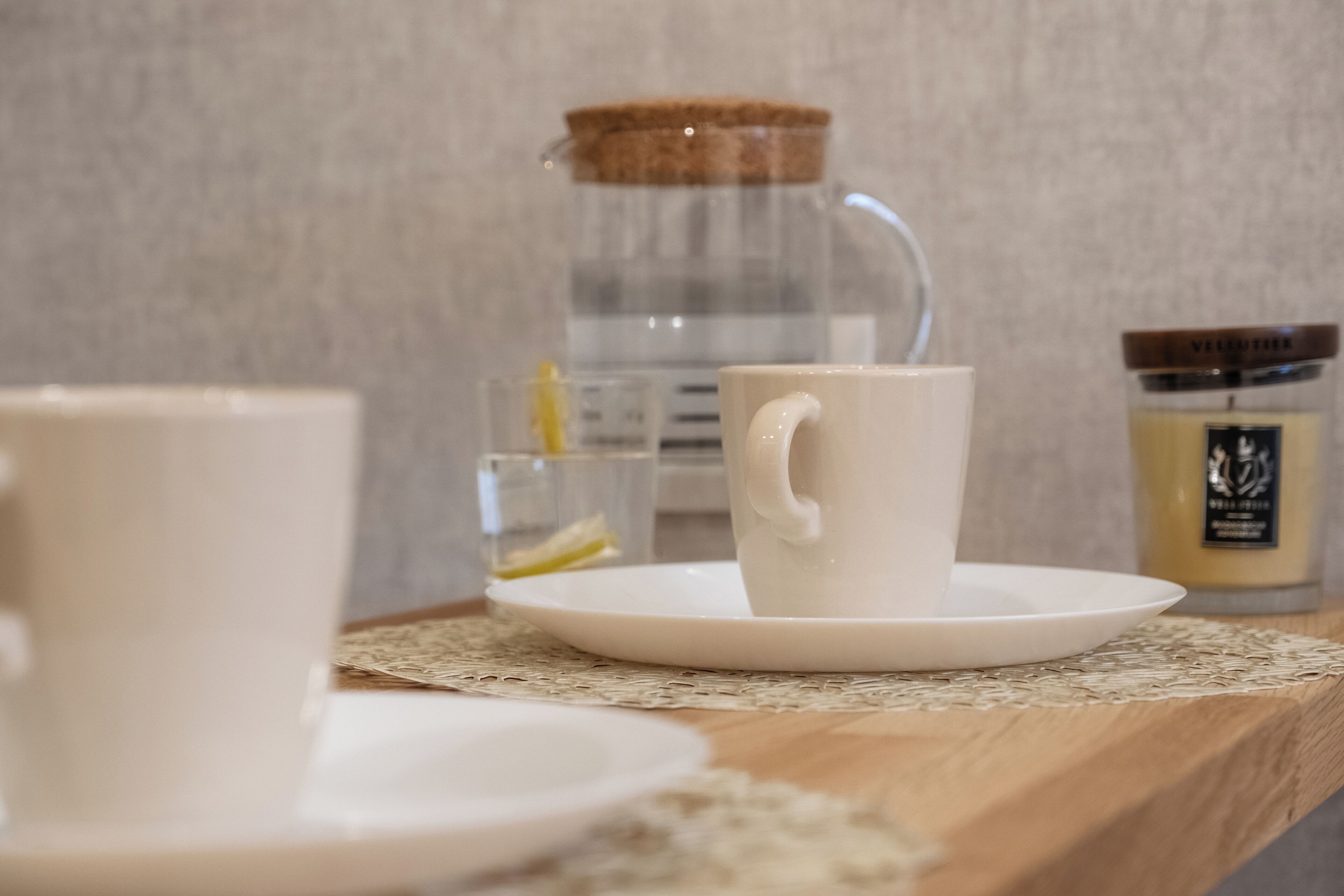 Une tasse blanche posée sur une assiette blanche, sur une table en bois, avec un verre d’eau et une bougie en arrière-plan.