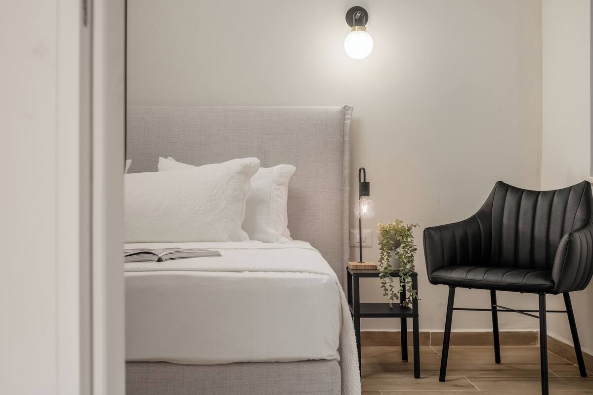 Quarto com cama, mesa de cabeceira, cadeira e luminária na parede.