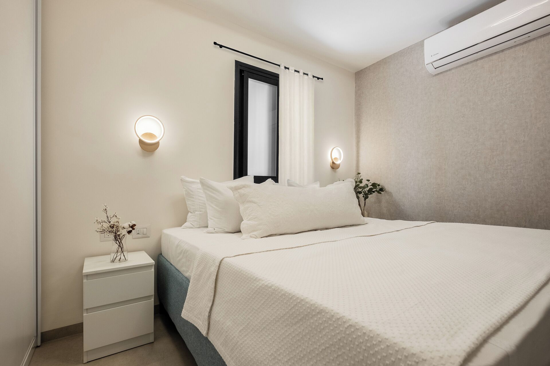 Un dormitorio con cama, mesitas de noche, luces fijadas a la pared, aire acondicionado y una ventana con cortinas.