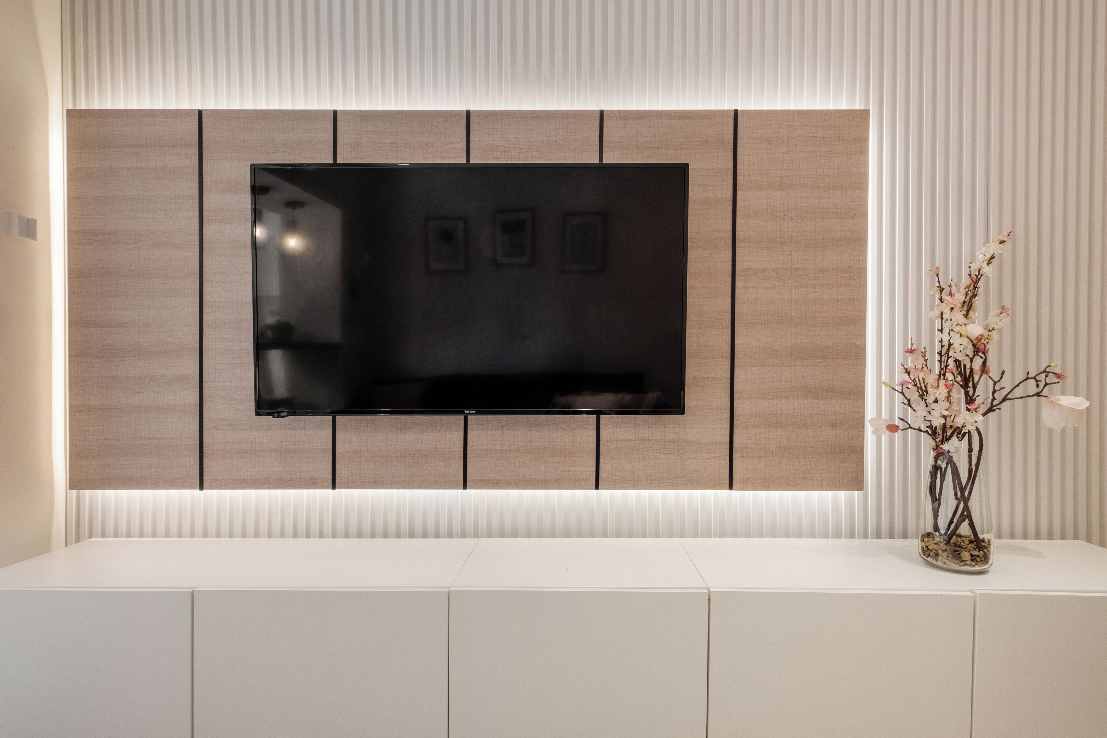 Una sala de estar moderna con un televisor de pantalla plana montado en una pared de paneles de madera, una consola blanca y un florero con flores.