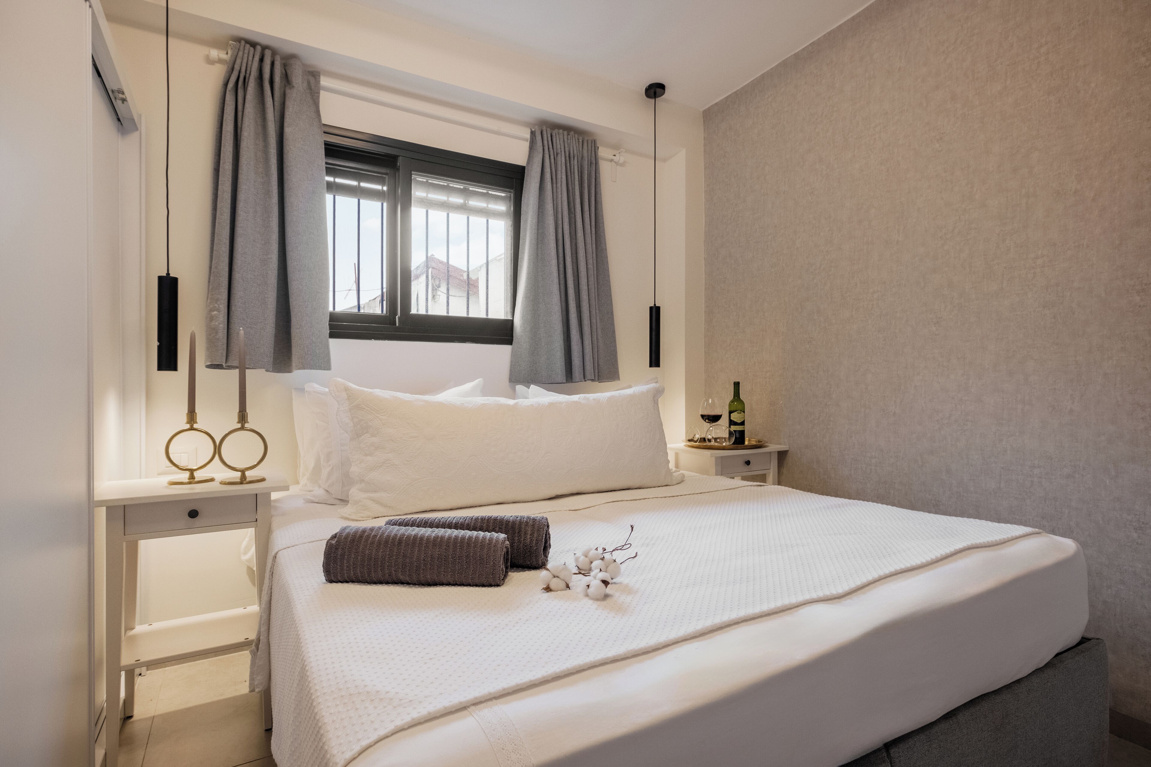Una cama bien hecha con una colcha blanca y almohadas grises, una mesita de noche con una botella de vino y una ventana con cortinas grises.
