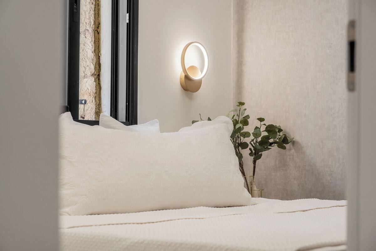 Uma cama bem arrumada com lençóis brancos, uma luminária fixada na parede e um vaso com planta.