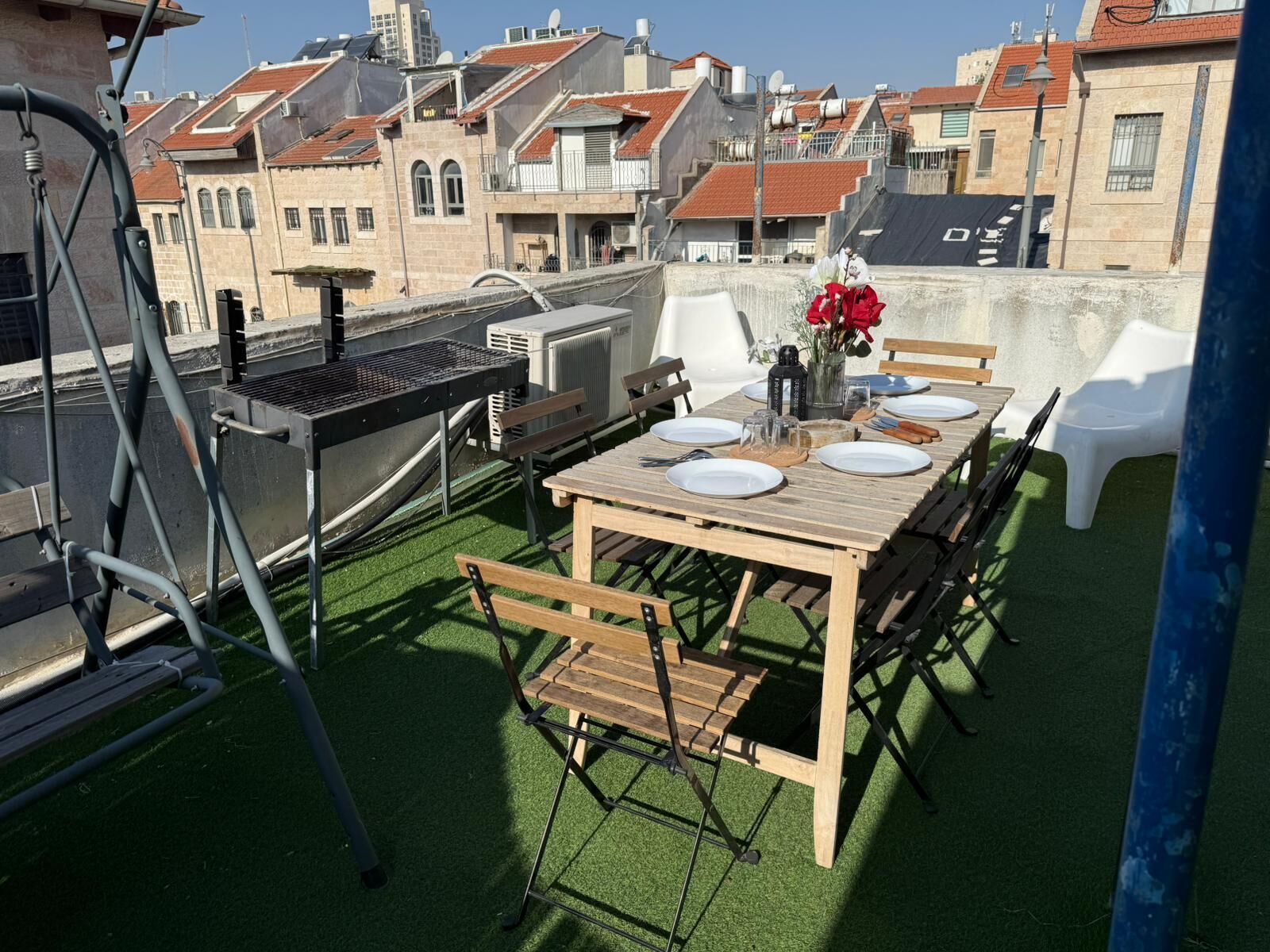 Un patio al aire libre con una mesa de madera dispuesta para cuatro personas, rodeado de sillas de plástico blanco.