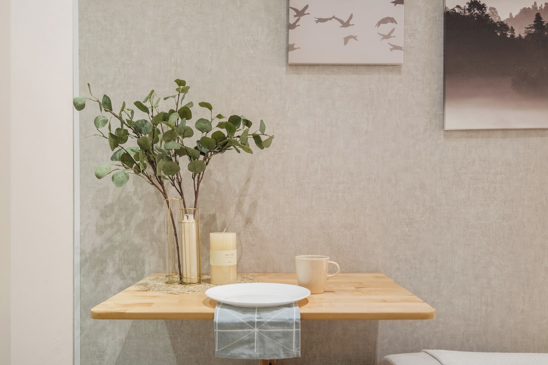 A wooden table with a vase of green leaves, a candle, and a white mug.
