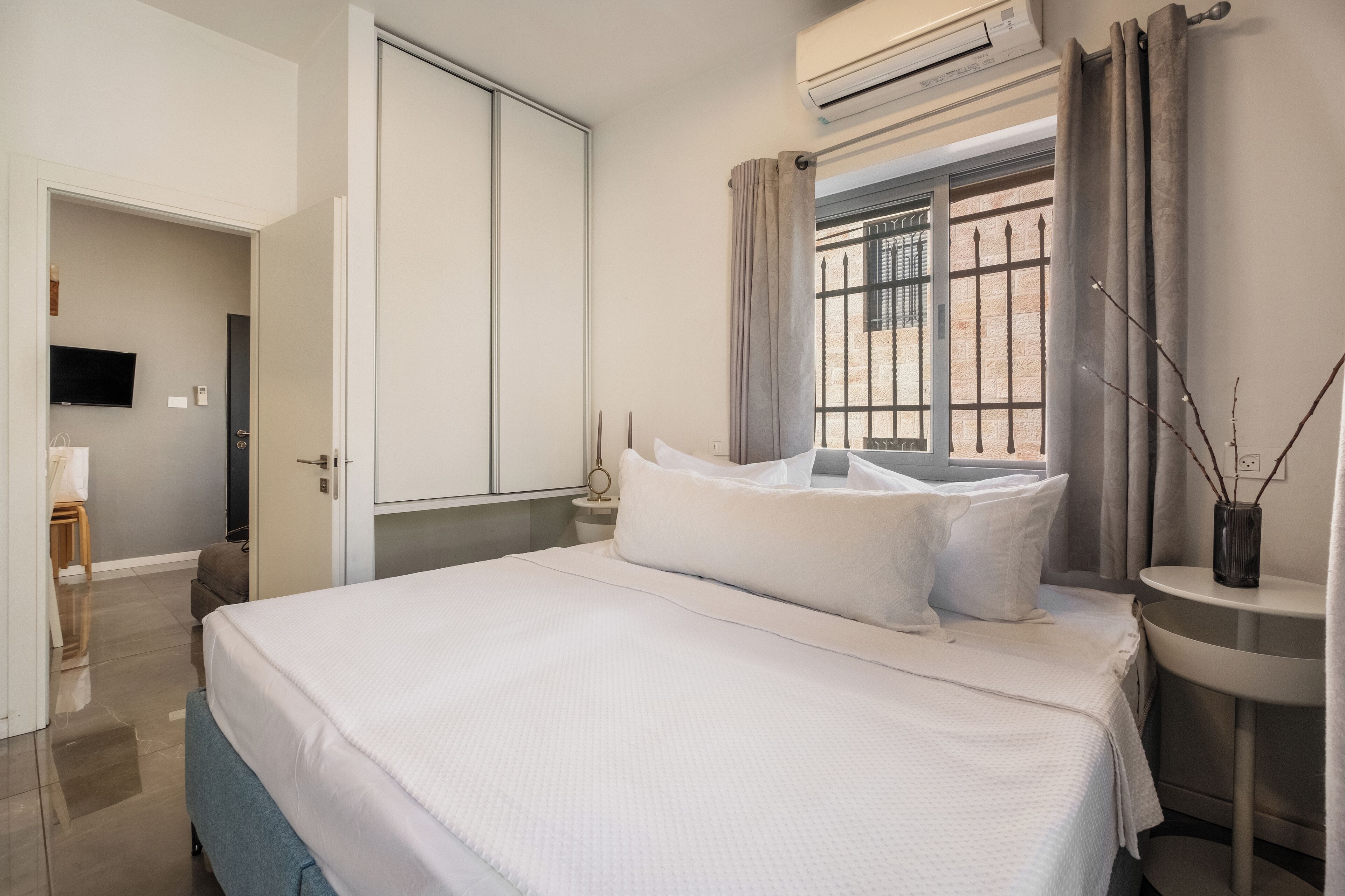 A hotel room with a bed, a TV, an air conditioner, a window with curtains, and a bedside table with a vase.