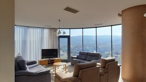 Ein modernes Wohnzimmer mit einem großen Flachbildfernseher, zwei Sofas, einem Couchtisch und Blick auf eine Stadtlandschaft durch große Fenster.