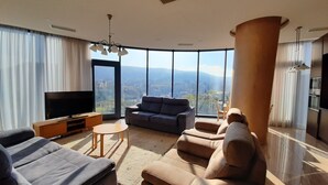 Ein modernes Wohnzimmer mit einem großen Flachbildfernseher, bequemen Sitzmöglichkeiten und Blick auf die Stadt durch große Fenster.