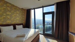 Ein Hotelzimmer mit einem großen Bett, Blick auf eine Stadtlandschaft durch eine Glastür und einem kleinen Tisch am Fenster.