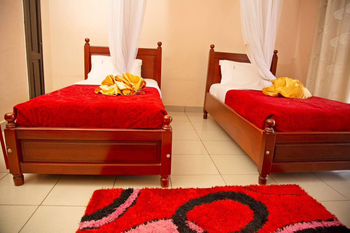 Two wooden beds with red bedding and white curtains, a red and black rug, and a white wall.