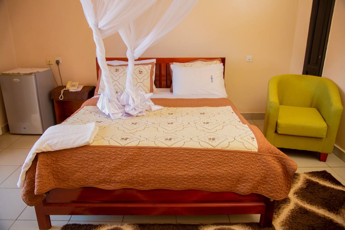 A hotel room with a bed, a nightstand, a telephone, a green chair, and a refrigerator.