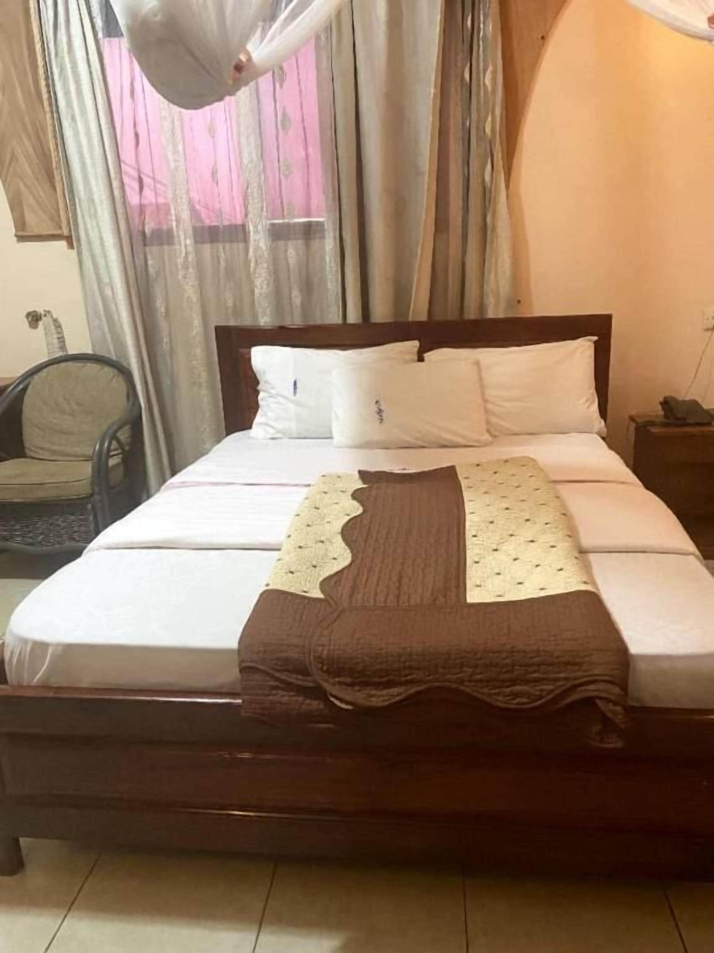 Een net opgemaakt bed met witte lakens en een bruine deken, een houten hoofdbord en een stoel op de achtergrond.