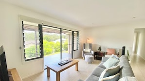 A spacious living room with a sofa, a coffee table, and a chair. There is a sliding glass door leading to a balcony with a view of greenery.