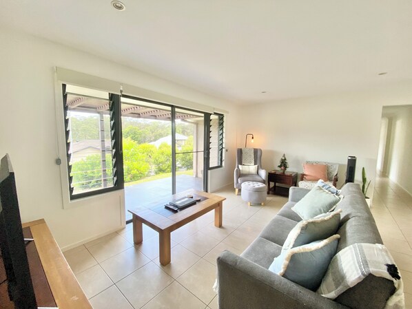 A spacious living room with a sofa, a coffee table, and a chair. There is a sliding glass door leading to a balcony with a view of greenery.