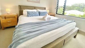 A neatly made bed with a striped blanket, two bedside tables with lamps, a painting on the wall, and a window with a view of greenery.
