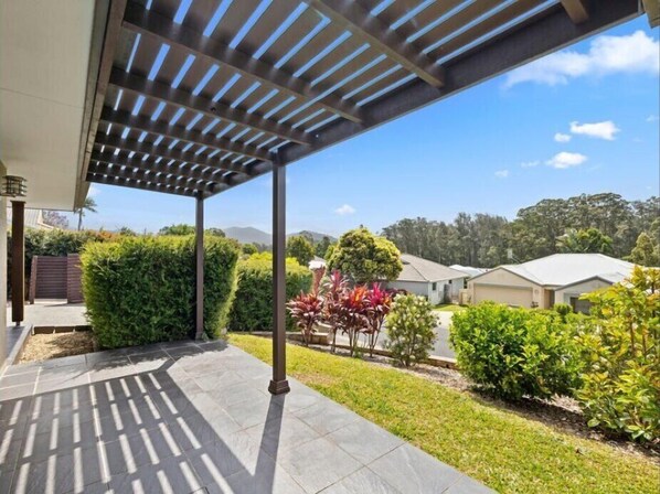 Un espacio exterior cubierto con un pérgola, suelo de piedra y jardines bien cuidados, que incluyen arbustos y césped.