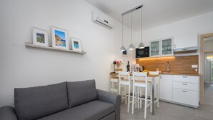 Un salon moderne avec un canapé gris, une petite table ronde et un coin cuisine doté de meubles blancs, d’un four à micro-ondes et de luminaires suspendus.
