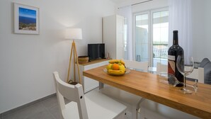 Un coin repas moderne avec une table en bois, un saladier de fruits, une bouteille de vin et un verre à vin. On y trouve également un téléviseur posé sur un meuble, une lampe et un tableau encadré accroché au mur.