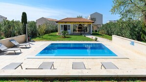 Une piscine avec des chaises longues, un patio couvert et une petite maison avec un toit en tuiles.