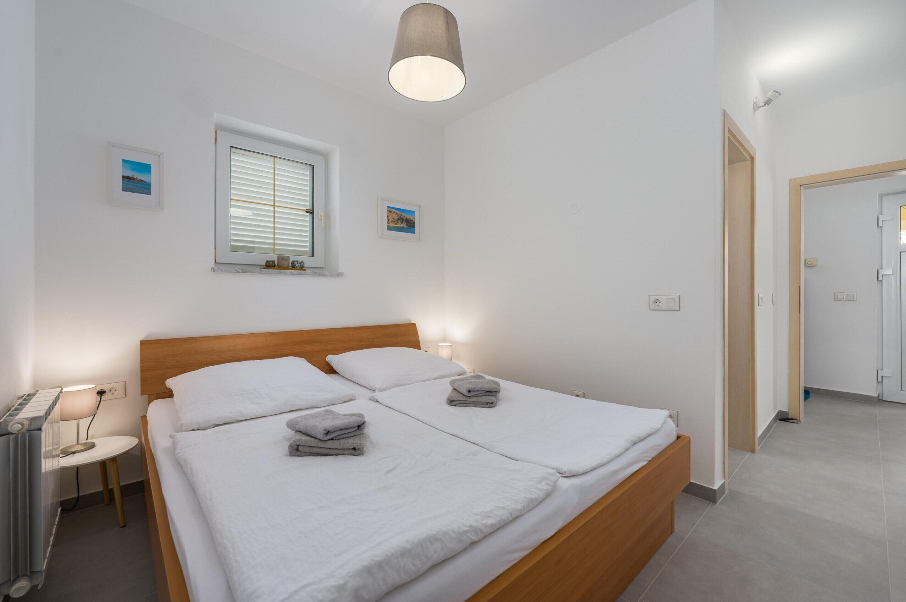 Un dormitorio limpio y moderno con una cama doble, lámparas de noche, un ventanal con persianas y una puerta que da a otra habitación.