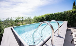 Una piscina rettangolare con ringhiere metalliche, circondata da un solarium in legno e vegetazione.