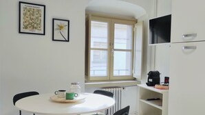 A small, clean room with a round table, four chairs, a window with blinds, a radiator, a shelf with a coffee maker, and a TV on the wall. - Conte Verde 8 by Wonderful Italy (Turin)