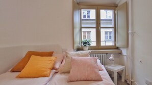 Ein Schlafzimmer mit Bett, Kissen, Nachttisch, Lampe und einem Fenster mit Blick auf Gebäude.