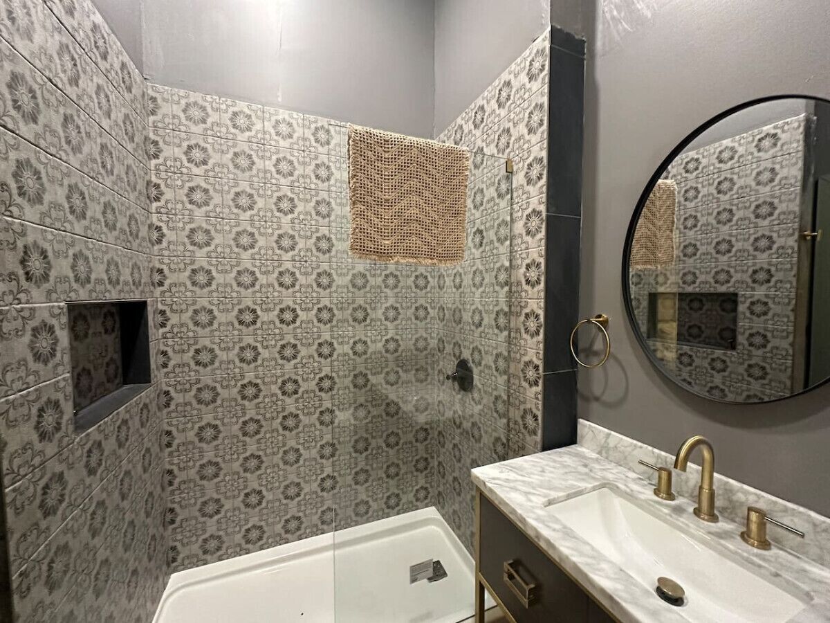 A bathroom with patterned tiles, a bathtub, a sink with gold fixtures, and a round mirror.